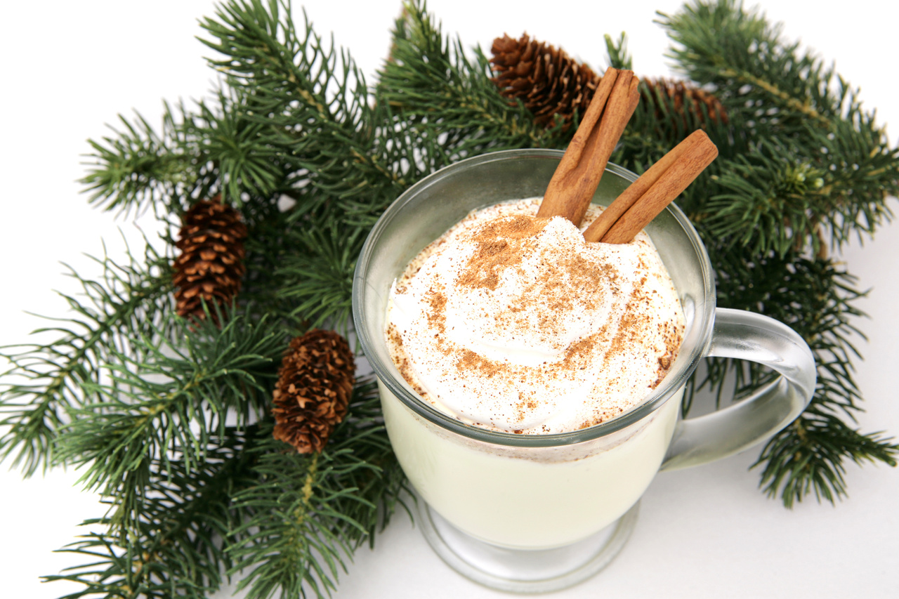 A mug of eggnog garnished with whipped cream, nutmeg and cinnamon sticks, nestled in pine branches. White background.